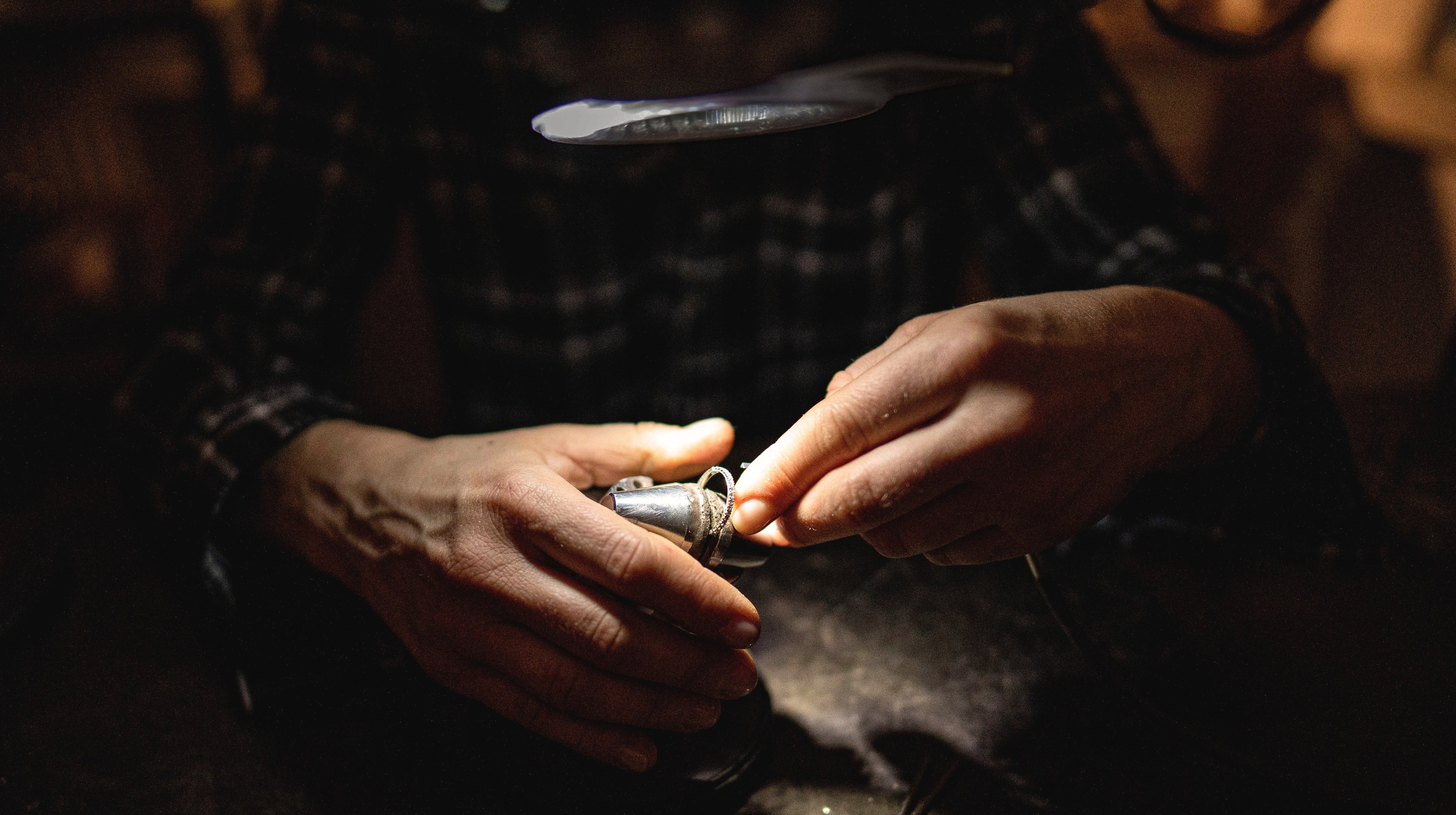 Close-up of hands working on a small metal object with a magnifying glass in a dimly lit setting from JYOCO aelier