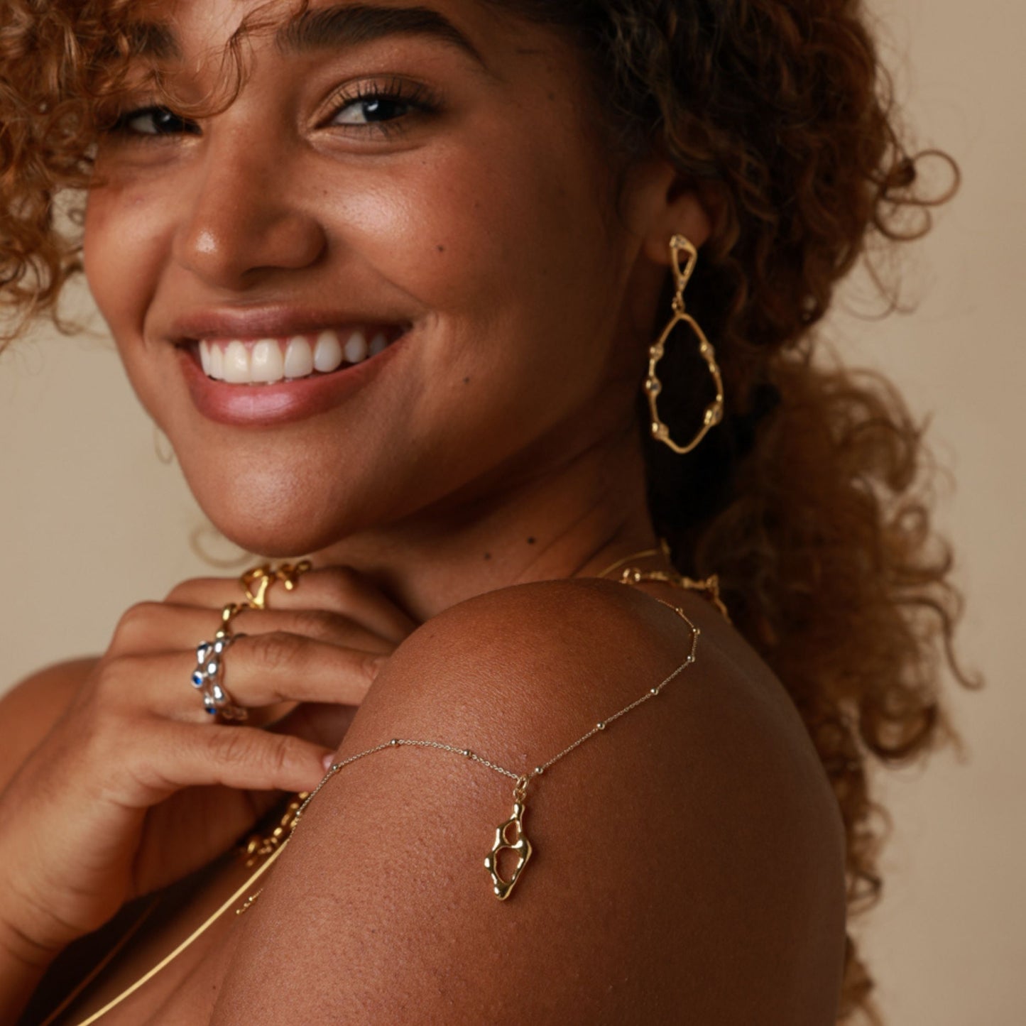 Woman wearing gold jewelry including earrings, necklace, and ring on a beige background